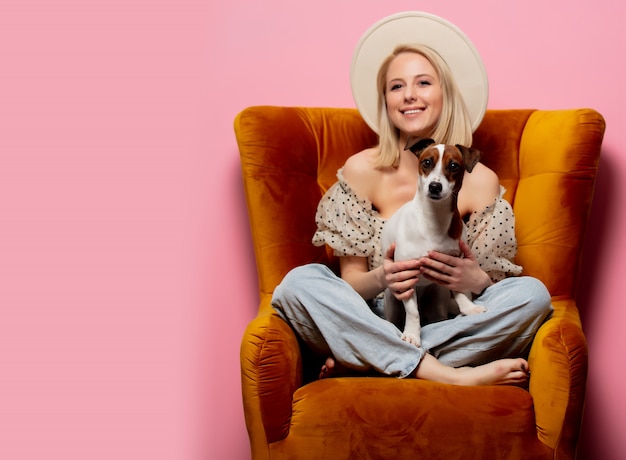 Hermosa mujer rubia con un perro en un sillón en la pared rosa