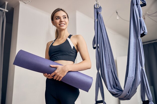 Hermosa mujer rubia en ropa deportiva de pie en el gimnasio cerca de las hamacas mientras sostiene la estera de yoga en las manos