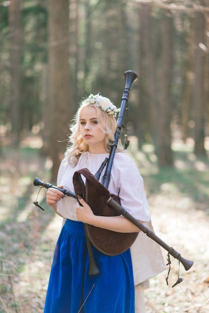 Hermosa mujer rubia con ropa eslava tocando la flauta y la pipa en el bosque de verano