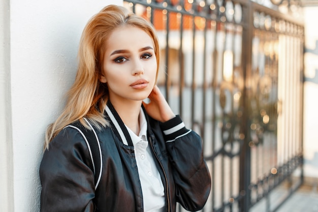 Hermosa mujer rubia en ropa de moda cerca de una pared blanca de una valla