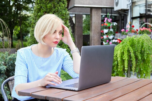 Hermosa mujer rubia sentada en la calle en un café y trabajando con una computadora portátil Lugar de trabajo remoto