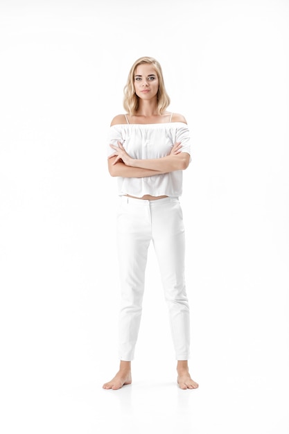 Hermosa mujer rubia seria en una blusa blanca y pantalones sobre un fondo blanco.
