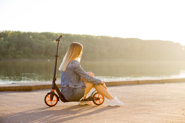 Hermosa mujer rubia sexy en ropa de mezclilla se sienta en su scooter en la calle frente al mar