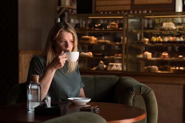 Hermosa mujer rubia se sienta en la cafetería y la panadería y bebe té o café y usa un teléfono inteligente