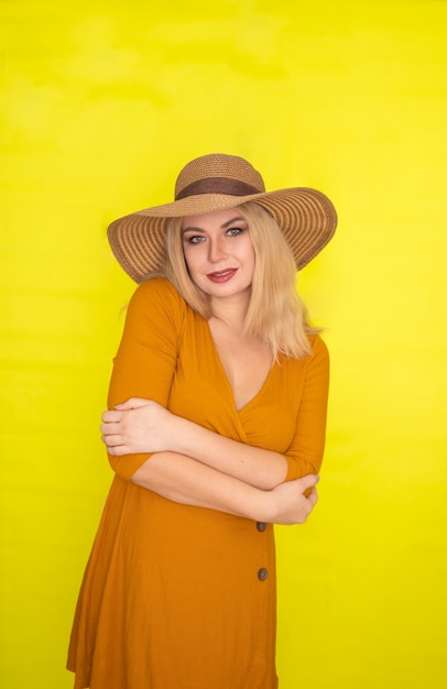 Hermosa mujer rubia con sombrero marrón y vestido amarillo oscuro posando sobre pared amarilla. Look de moda de verano
