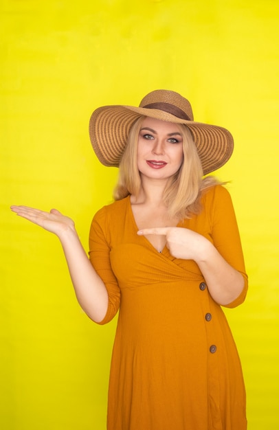 Hermosa mujer rubia con sombrero marrón y vestido amarillo oscuro posando sobre pared amarilla. Look de moda de verano