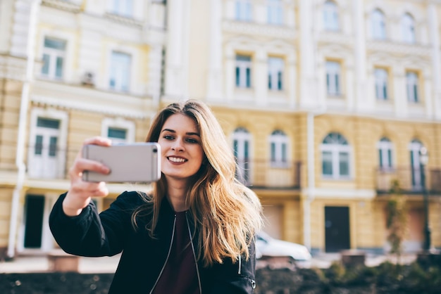 Hermosa mujer rubia sonriente haciendo clic en selfie usando un teléfono inteligente moderno en la calle de la ciudad