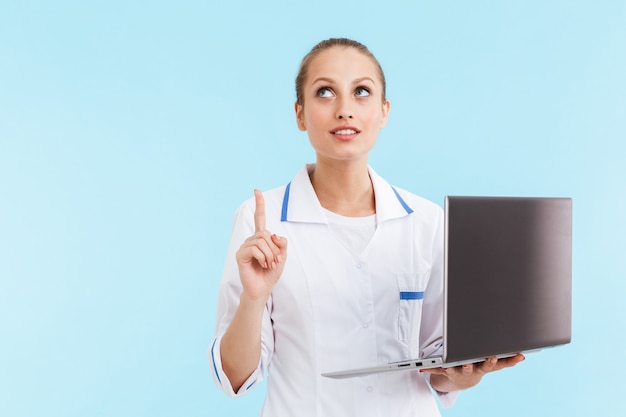 Hermosa mujer rubia sonriente médico vistiendo uniforme que se encuentran aisladas sobre la pared azul, sosteniendo la computadora portátil, apuntando con el dedo hacia arriba