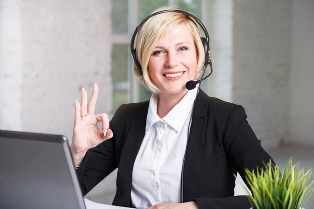 Hermosa mujer rubia sonriente operadora del centro de llamadas vestida con un elegante traje negro que muestra el signo de ok en la mesa con la computadora portátil