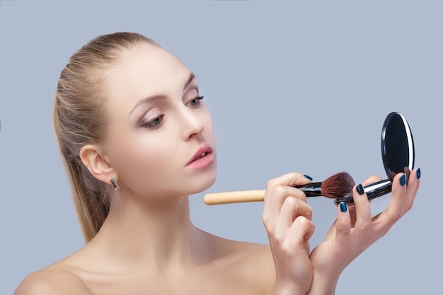 hermosa mujer rubia sosteniendo pincel de maquillaje y mirando en el espejo sobre un fondo gris. de cerca