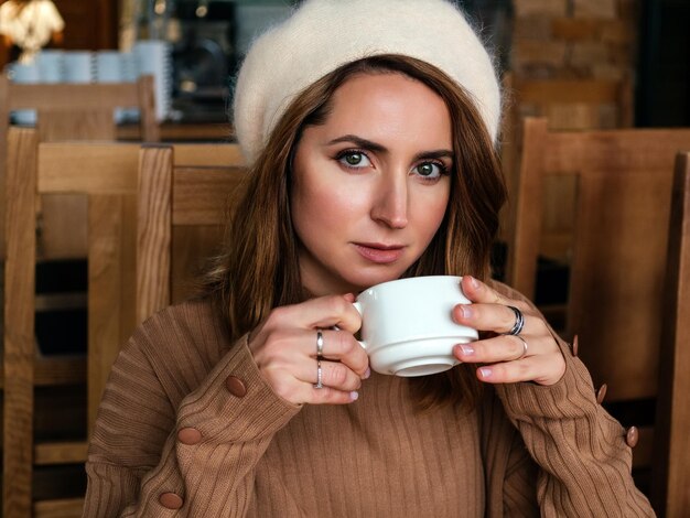 hermosa mujer rubia con suéter beige y boina bebe de una taza en un café retrato ojos expresivos