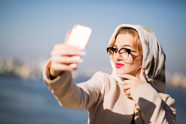 Hermosa mujer rubia tomando selfie en teléfonos inteligentes. Mujer, viste, chal, y, anteojos