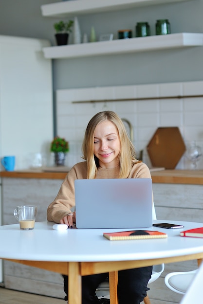 Hermosa mujer rubia trabajando con su computadora portátil mientras está sentado en la cocina de su apartamento