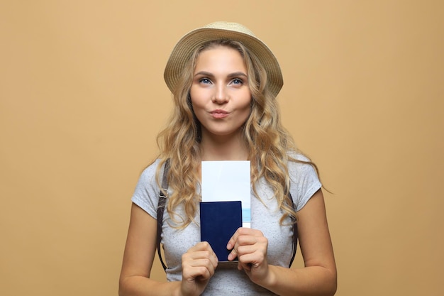 Hermosa mujer rubia vestida con ropa de verano posando con pasaporte con entradas sobre fondo beige.