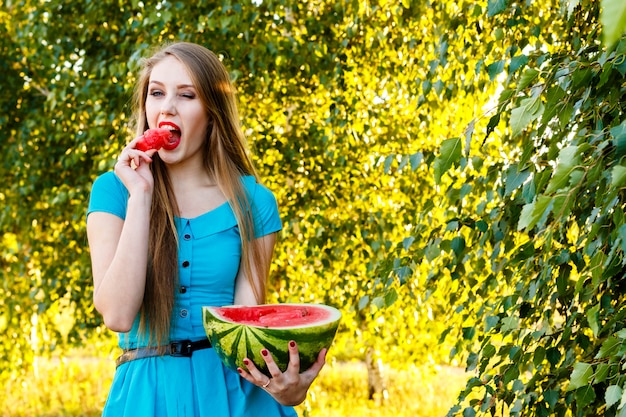 Foto hermosa mujer rubia en vestido azul comiendo una sandía al aire libre