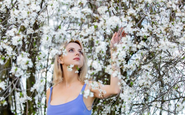 Hermosa mujer rubia en vestido azul junto al árbol floreciente