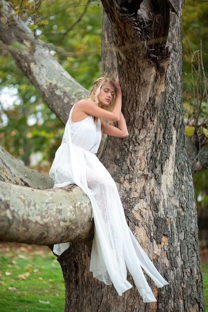 Hermosa mujer rubia con un vestido blanco sentada en un árbol