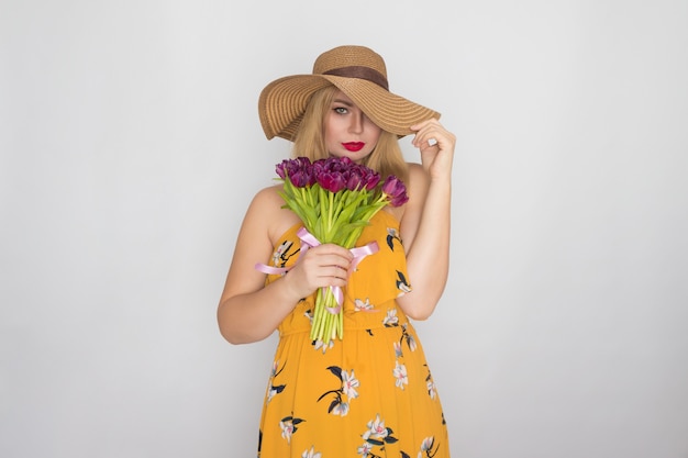 Hermosa mujer rubia en vestido floral amarillo y sombrero de paja con ramo de tulipanes púrpuras