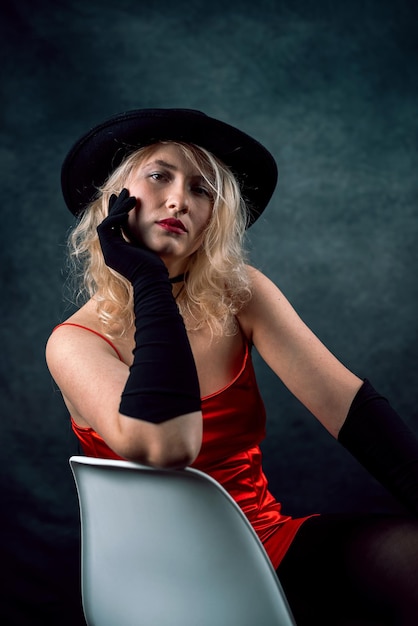 Hermosa mujer rubia en vestido de noche elegante rojo y pose de sombrero negro en estudio aislado sobre fondo oscuro Concepto de estilo de moda