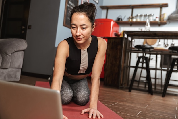 Hermosa mujer sana haciendo ejercicios de yoga mientras está sentado en una colchoneta de fitness en casa, usando una computadora portátil, estirando