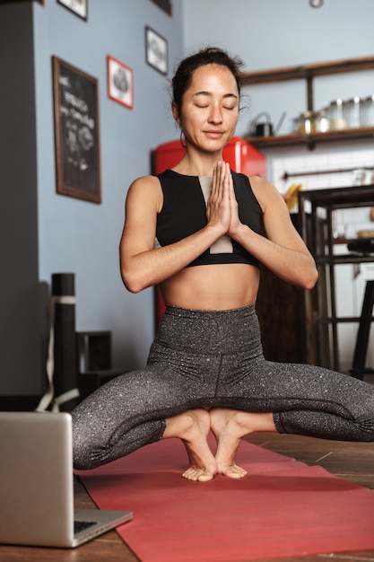 Hermosa mujer sana haciendo ejercicios de yoga mientras está sentado en una colchoneta de fitness en casa, usando una computadora portátil, estirando