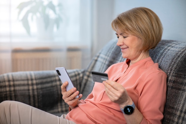 Hermosa mujer senior anciana positiva feliz en casa con teléfono móvil, smartphone, compra, uso, sosteniendo en la mano tarjeta de crédito para compras por internet, sonriendo. Concepto de pago online.