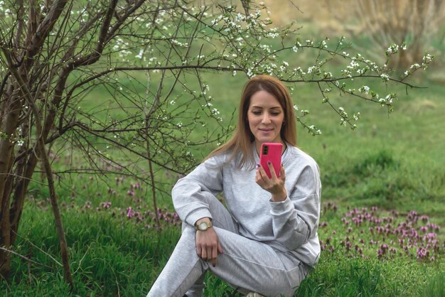 hermosa mujer sentada en el suelo en un parque de primavera esperando a alguien y mirando su teléfono