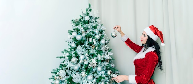 Hermosa mujer en un sexy disfraz de Santa Claus decora las decoraciones del árbol de Navidad en casa. Niña cuelga una bola plateada en un árbol de Navidad verde. Preparativos para la celebración del año nuevo.