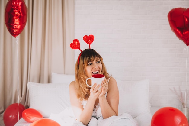 Una hermosa mujer se sienta en la cama y sostiene una taza blanca en sus manos. día de San Valentín