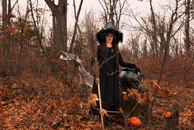 Hermosa mujer con sombrero de bruja y traje sosteniendo escoba cerca del caldero en el bosque de otoño Concepto de Halloween Enfoque selectivo