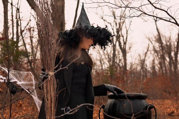 Hermosa mujer con sombrero de bruja y traje sosteniendo escoba cerca del caldero en el bosque de otoño Concepto de Halloween Enfoque selectivo