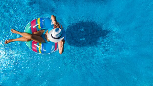 Hermosa mujer con sombrero en la piscina vista aérea desde arriba, jovencita en bikini se relaja y nada en anillo inflable donut y se divierte en el agua