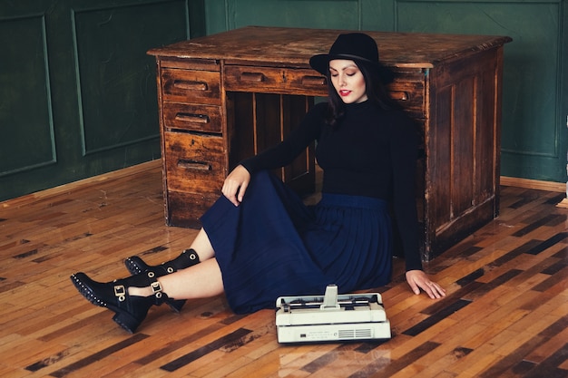 Hermosa mujer con sombrero se sienta junto a la mesa de roble con máquina de escribir vintage.