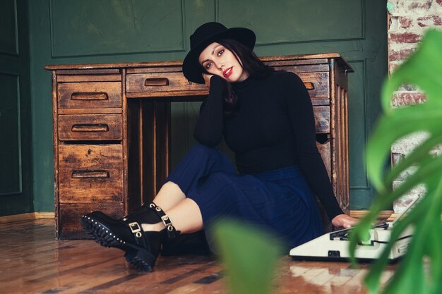 Hermosa mujer con sombrero se sienta junto a la mesa de roble con máquina de escribir vintage.