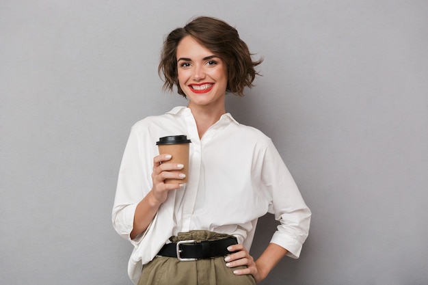Hermosa mujer sonriendo y sosteniendo café para llevar, aislado sobre pared gris