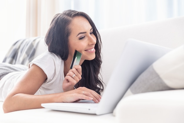 Hermosa mujer sonriente acostada en un sofá blanco de compras en línea, tocando la computadora portátil con la mano derecha mientras sostiene una tarjeta de crédito verde en la mano izquierda.