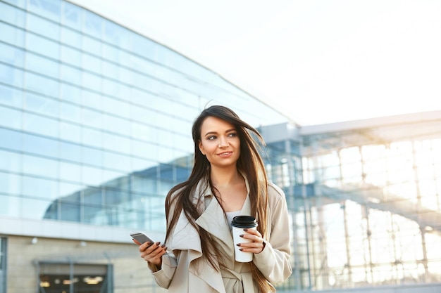 Hermosa mujer sonriente caminando cerca de la oficina desde el trabajo con una taza de café y enviando mensajes de texto por teléfono móvil