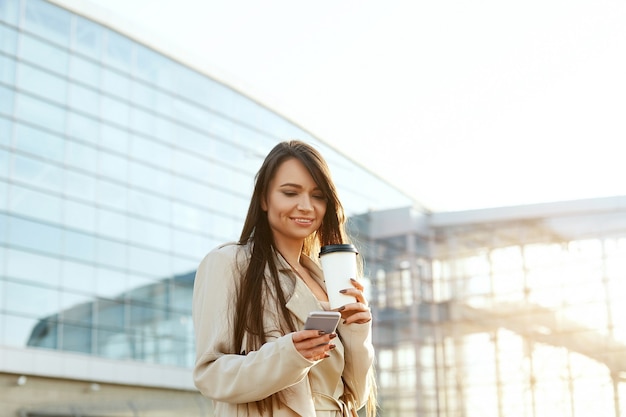 Hermosa mujer sonriente caminando cerca de la oficina del trabajo con taza de café y mensajes de texto en el teléfono móvil. Comunicación telefónica.