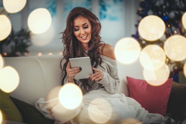 Hermosa mujer sonriente está usando una tableta y sentada en un sofá envuelto en una manta en la víspera de Navidad.