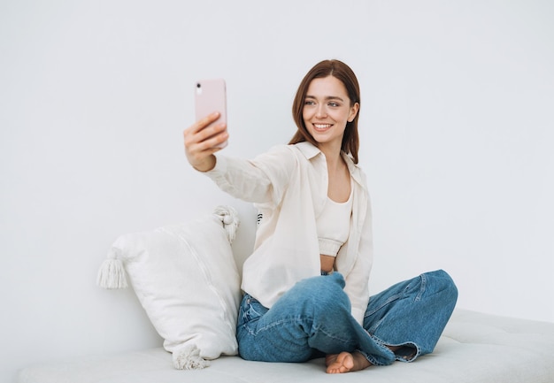 Hermosa mujer sonriente estudiante adolescente con cabello largo oscuro en camisa blanca tomando selfie en el teléfono móvil en la mano sentada en el sofá en casa