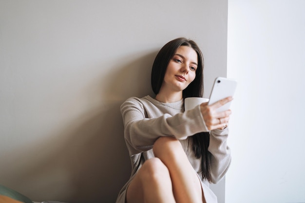 Hermosa mujer sonriente estudiante adolescente con cabello largo oscuro en ropa de casa con una taza de café o té por la mañana usando medios cosiales de teléfonos móviles en el fondo gris