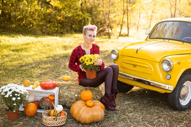 Hermosa mujer sonriente con flores y calabazas en el fondo del parque de otoño cerca del auto amarillo