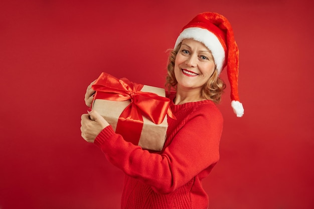 Hermosa mujer sonriente en un gorro de Navidad con un regalo en sus manos posando sobre un fondo rojo.