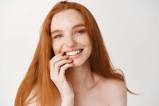 Hermosa mujer sonriente con largo cabello rojo natural y dientes blancos mirando feliz al frente, de pie desnuda en la pared blanca