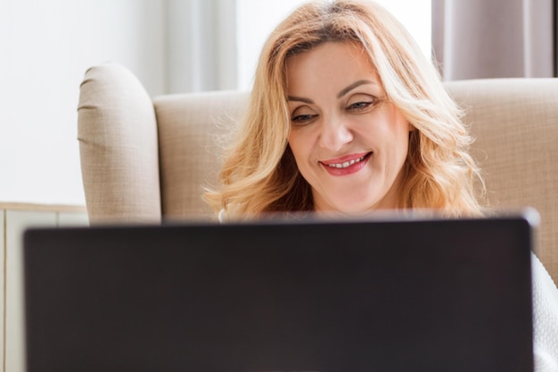 Hermosa mujer sonriente madura trabajando en una laptop y sentada en una silla grande y cómoda en casa