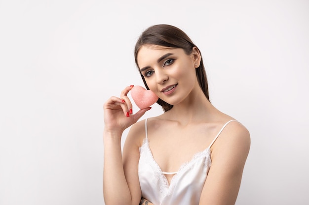 Hermosa mujer sonriente con maquillaje natural de piel limpia y dientes blancos sobre un fondo gris Sosteniendo un corazón higiénico rosa