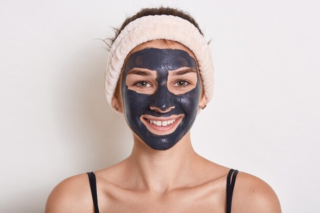 Hermosa mujer sonriente con máscara facial de arcilla negra en la cara, mirando a la cámara con expresión feliz, con diadema en la cabeza, posando aislada sobre fondo blanco.