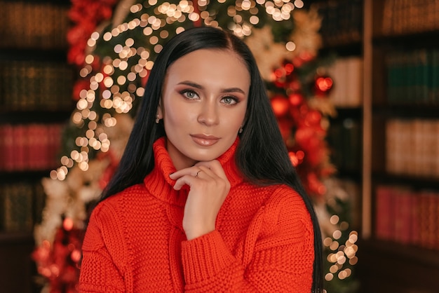 Hermosa mujer sonriente modelo Maquillaje peinado largo saludable Elegante dama con un suéter rojo en el contexto de las luces del árbol de Navidad feliz año nuevo