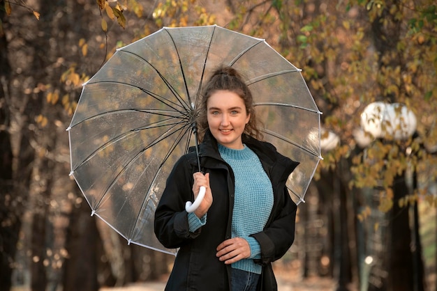 Hermosa mujer sonriente con ojos azules mira a la cámara Caminar en el parque de otoño con paraguas transparente