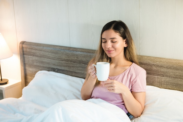 Hermosa mujer sonriente con los ojos cerrados sosteniendo una taza mientras está acostado en la cama con ropa de cama blanca